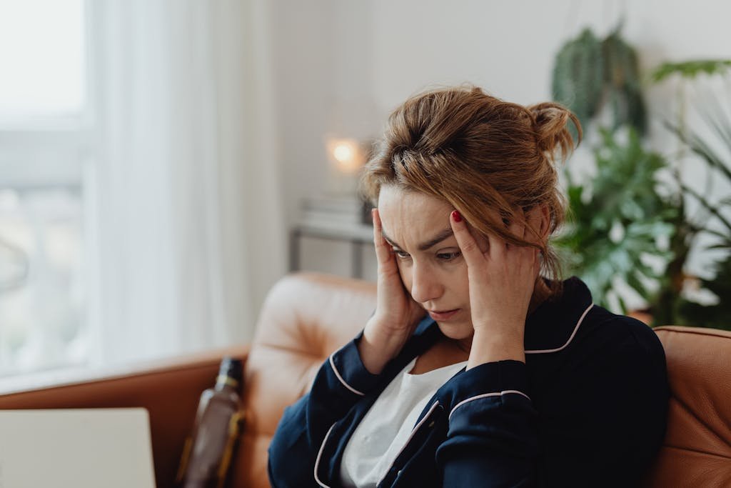 A woman in pajamas sits indoors with hands on head, showing stress and frustration.